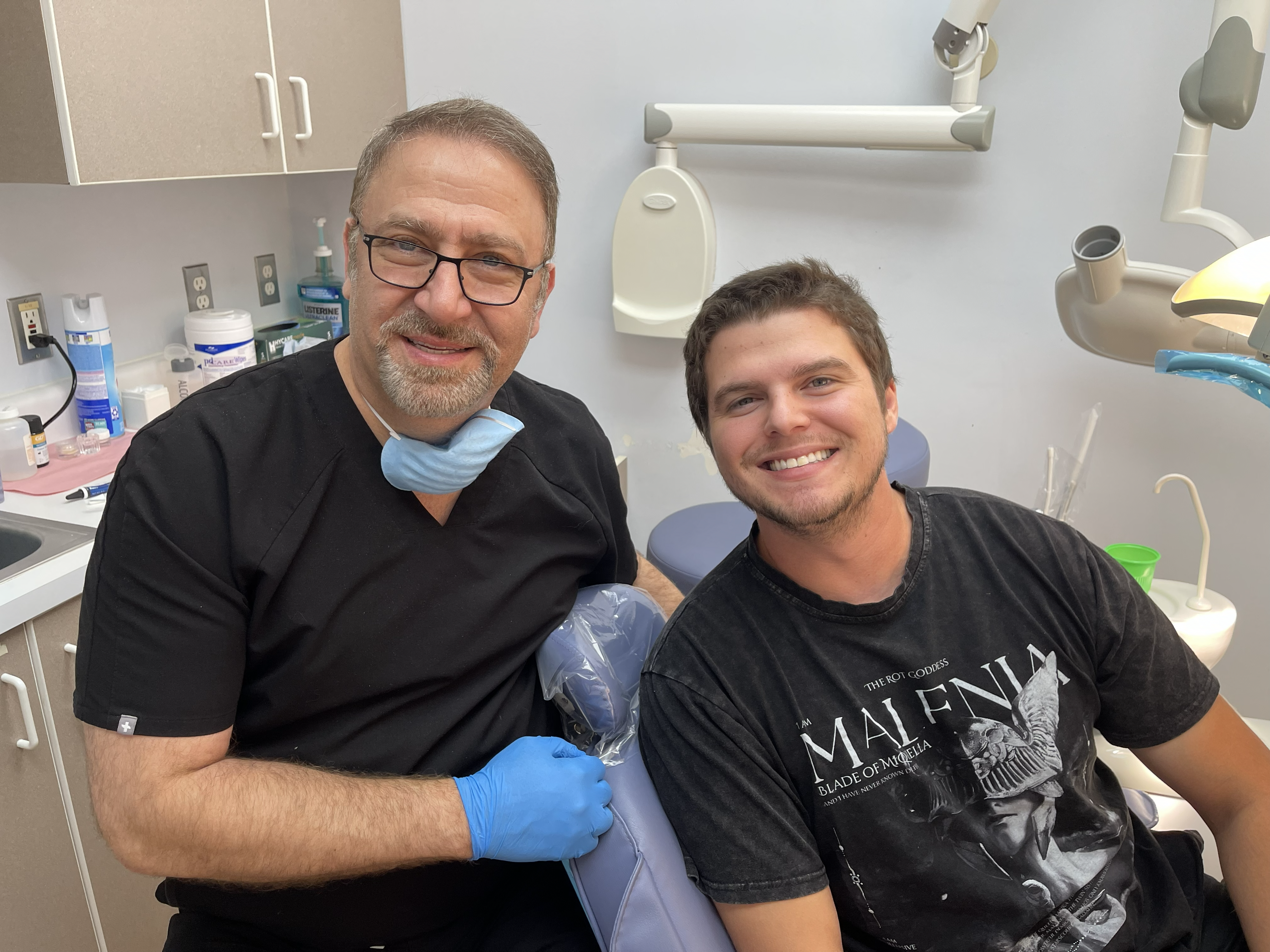 A dentist in teal scrubs speaking with a relaxed patient in a bright treatment room